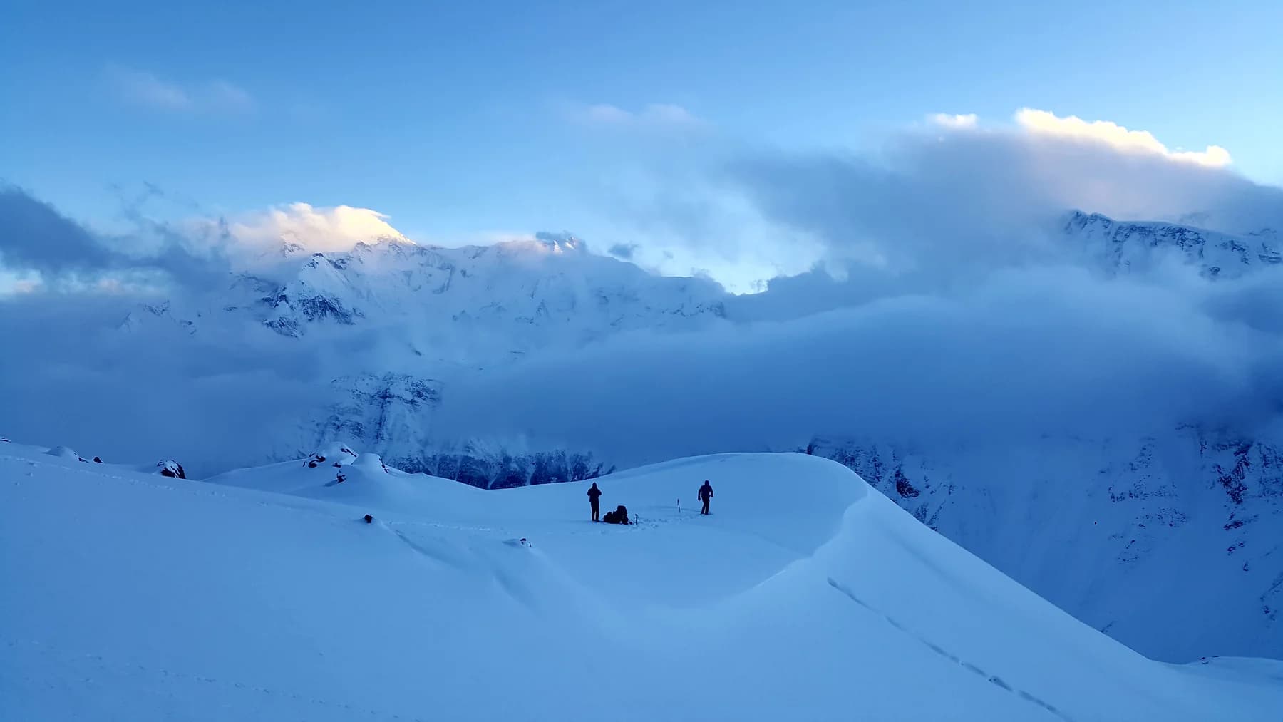 Two small figures on a snow-covered ridge beneath cloud-wrapped mountains in blue light