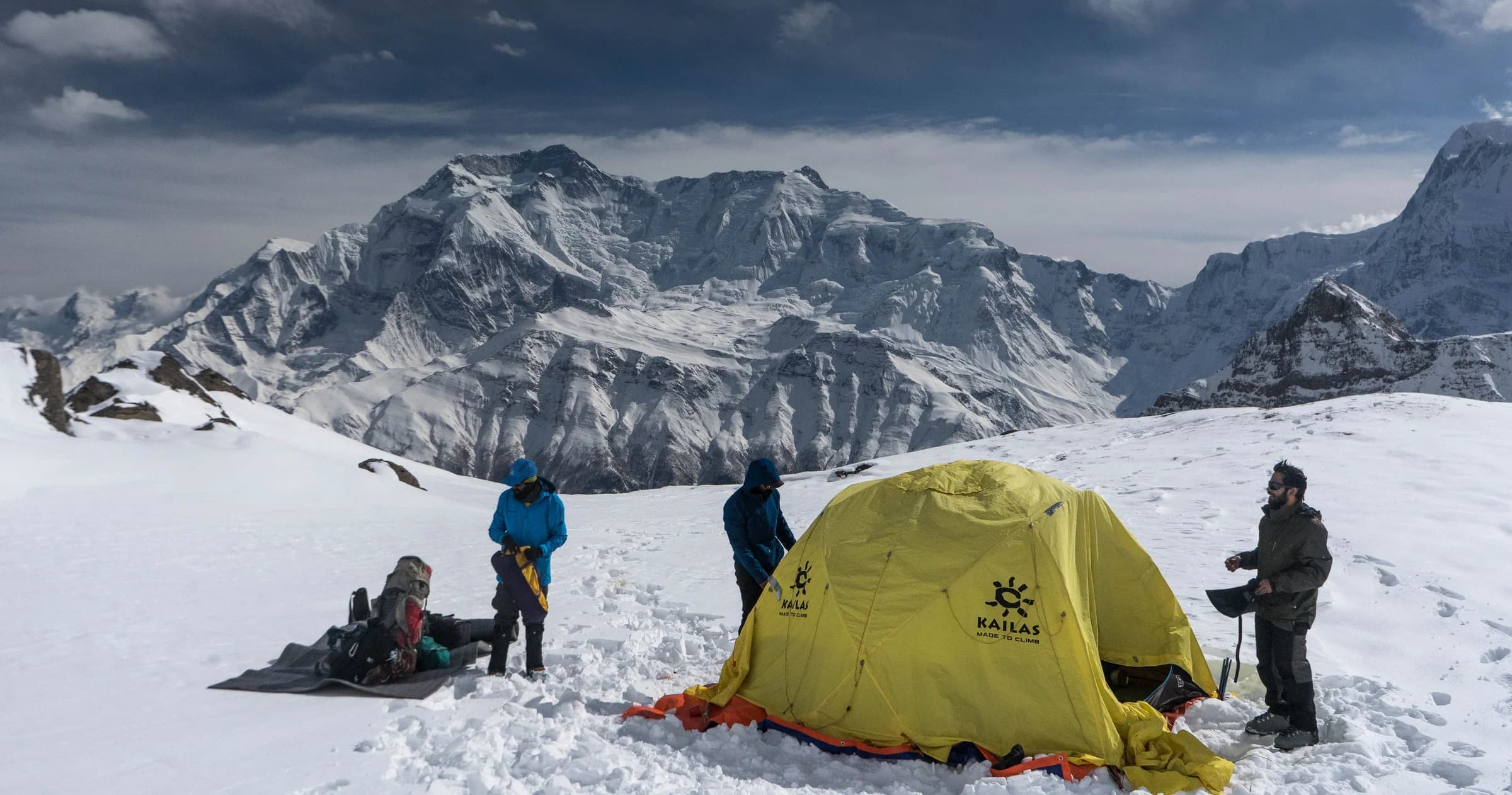 A yellow expedition tent and climbers on a snowy campsite beneath a Himalayan mountain wall