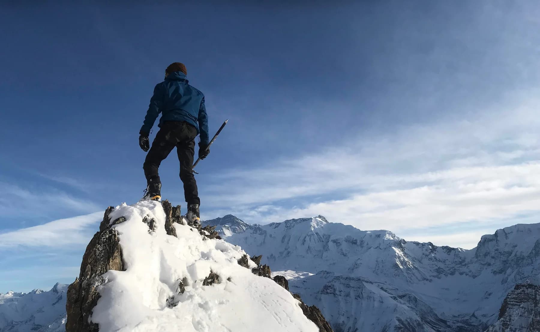 Intiaz Mahmud holding an ice axe on a snow-covered rocky ridge above a mountain range