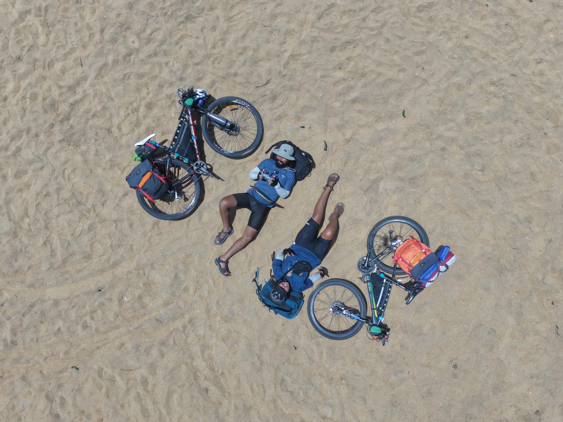 An overhead view of Intiaz and Subah resting on sand between their loaded bicycles