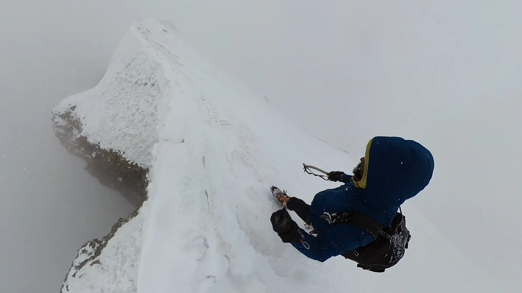 A solo climber traverses an exposed snow ridge in cloud and falling snow