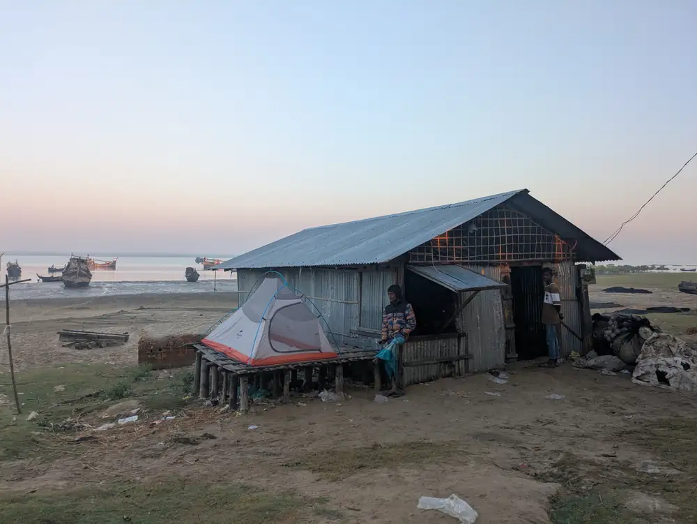 A tent on a raised wooden platform beside a tin-roofed riverside building