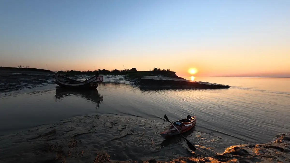 An orange kayak and a wooden boat beside a sandy riverbank at sunset