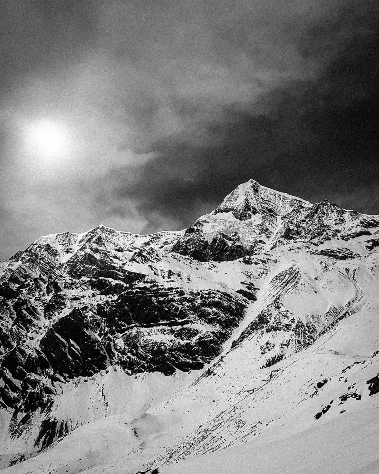 A black-and-white mountain photograph with snow-filled gullies, jagged ridges and bright sun through cloud